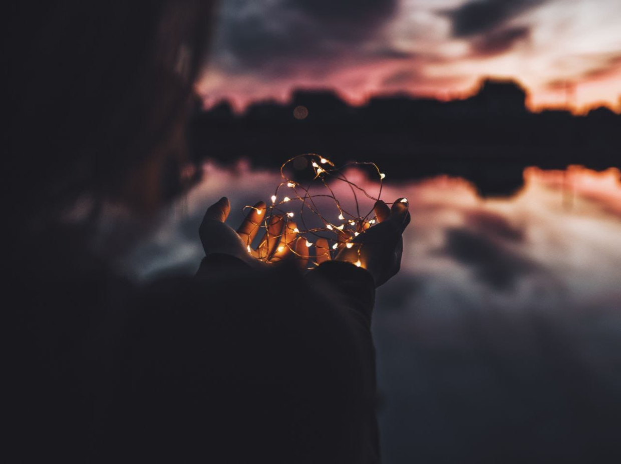 Hände halten wie in einer Schale eine Lichterkette. Im Mittelgrund ein See in dem sich im Abendlicht