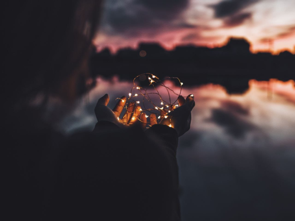 Hände halten wie in einer Schale eine Lichterkette. Im Mittelgrund ein See in dem sich im Abendlicht