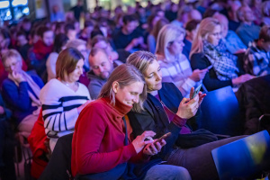 Zuschauer:innen in einem voll besetzten Veranstaltungsraum, viele mit Blick auf ihre Smartphones. Im Vordergrund lachen zwei Frauen, während sie gemeinsam auf ihre Handys schauen. Die Szene ist bunt beleuchtet und zeigt ein konzentriertes, interaktives Publikum.          ChatGPT fragen