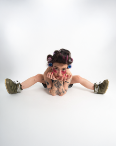 Eine junge Frau mit dunklen Haaren und Lockenwicklern sitzt in einer extrem flexiblen Pose auf dem Boden. Ihr Gesicht ruht auf ihren Händen, während ihre Beine weit auseinander gespreizt sind. Sie trägt olivgrüne Moon Boots und hat auffälliges Make-up mit roten Akzenten unter den Augen. Ihre Arme sind mit zahlreichen Tattoos verziert. Der Hintergrund ist schlicht und weiß, wodurch der Fokus vollständig auf ihr ungewöhnliches Posing und ihren ausdrucksstarken Blick gelenkt wird.