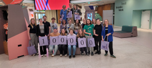 Gruppenfoto von rund 20 Personen, die im Innenraum eines modernen Gebäudes sitzen und stehen. Sie halten violette Taschen hoch, auf denen zusammen die Zahl „100000“ steht.